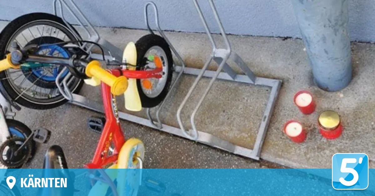 Ein rotes Dreirad mit gelben Rädern ist teilweise an einem Metall-Fahrradständer befestigt, möglicherweise mit einem Schloss gesichert. In der Nähe sind zwei rote Kerzen.