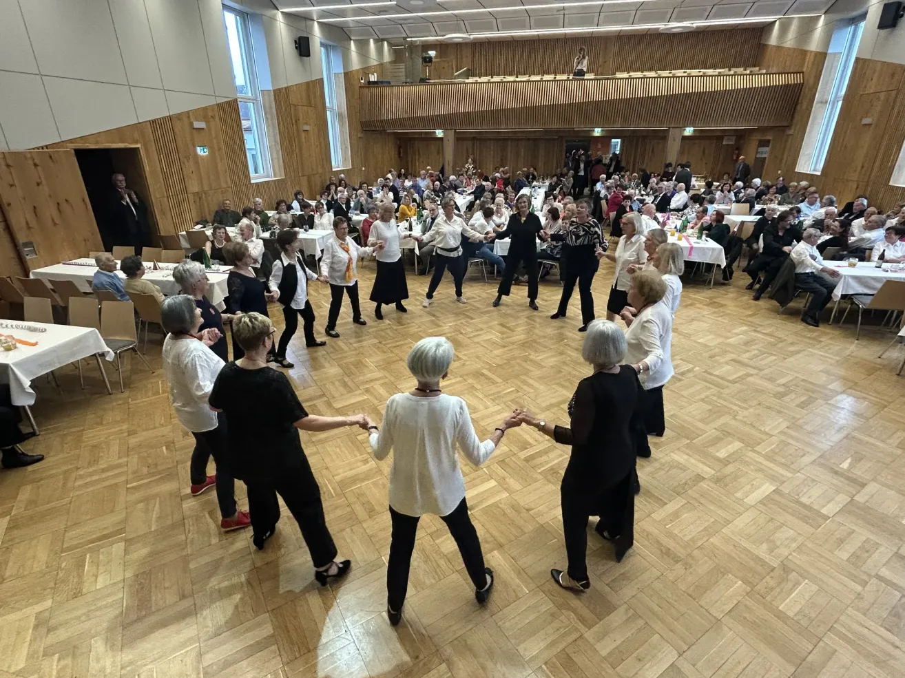 Eine Gruppe älterer Frauen tanzt in einem großen Raum mit Holzböden in einem Kreis. Zuschauer sitzen an Tischen am Rand.