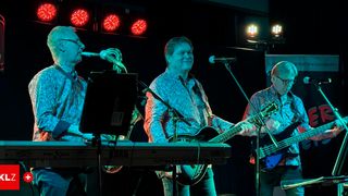 Drei Männer auf einer Bühne. Einer spielt Saxophon, ein anderer spielt Klavier und der dritte spielt Gitarre. Sie werden von roten Bühnenlichtern beleuchtet.