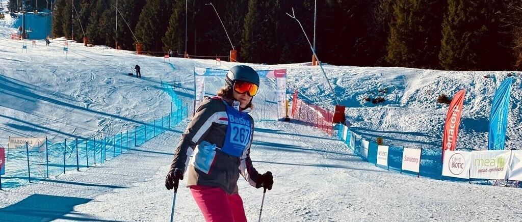 Ein Skifahrer steht auf einem schneebedeckten Hang mit der Nummer 267 auf ihrer Jacke. Sie trägt einen Helm und eine Schutzbrille. Es gibt Barrieren und Schilder am Hang.