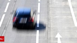 Ein dunkelblaues Fahrzeug fährt auf einer verschwommenen Straße mit weißen Markierungen und Pfeilen. Das Bild ist unscharf.