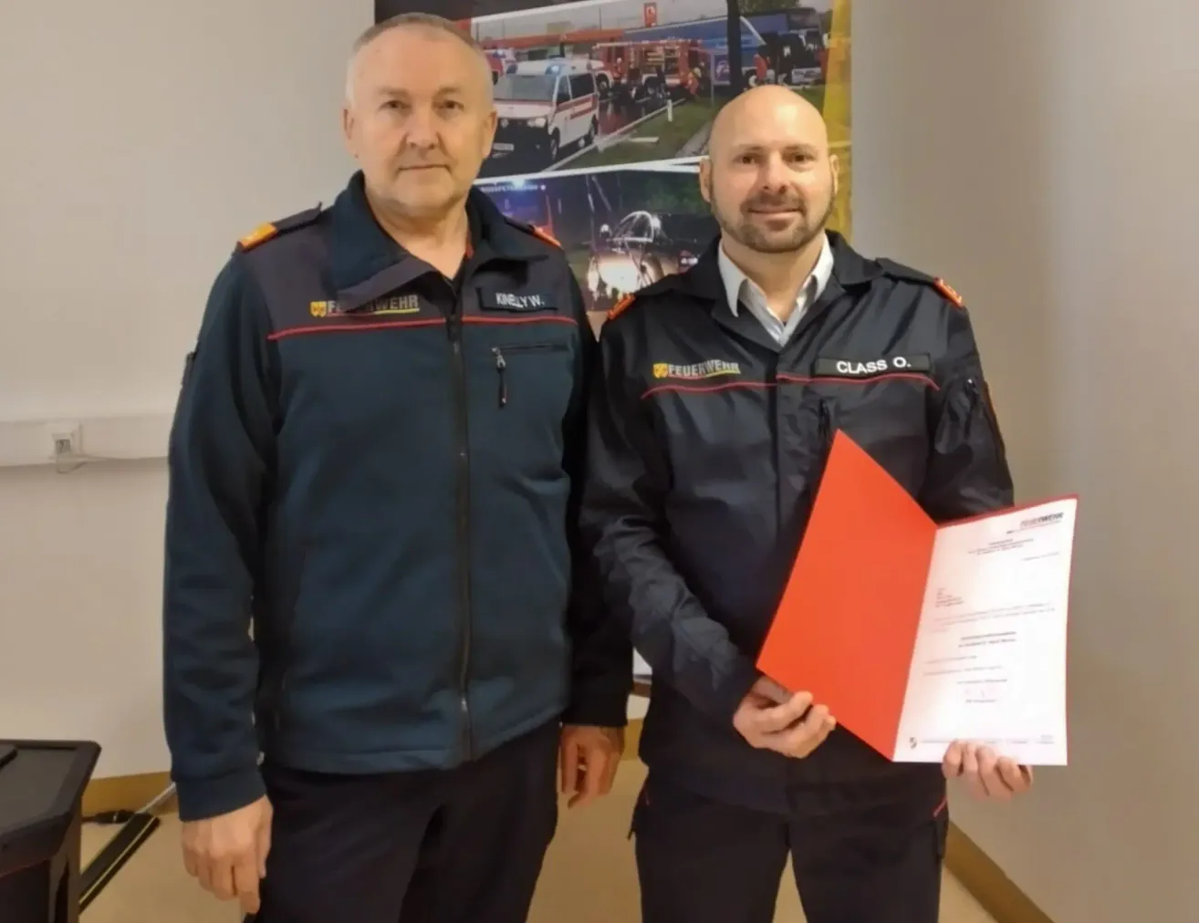Zwei Feuerwehrleute in Uniform, einer hält ein Zertifikat, stehen neben einem Poster mit Rettungsfahrzeugen.