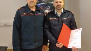 Zwei Feuerwehrleute in Uniform, einer hält ein Zertifikat, stehen neben einem Poster mit Rettungsfahrzeugen.