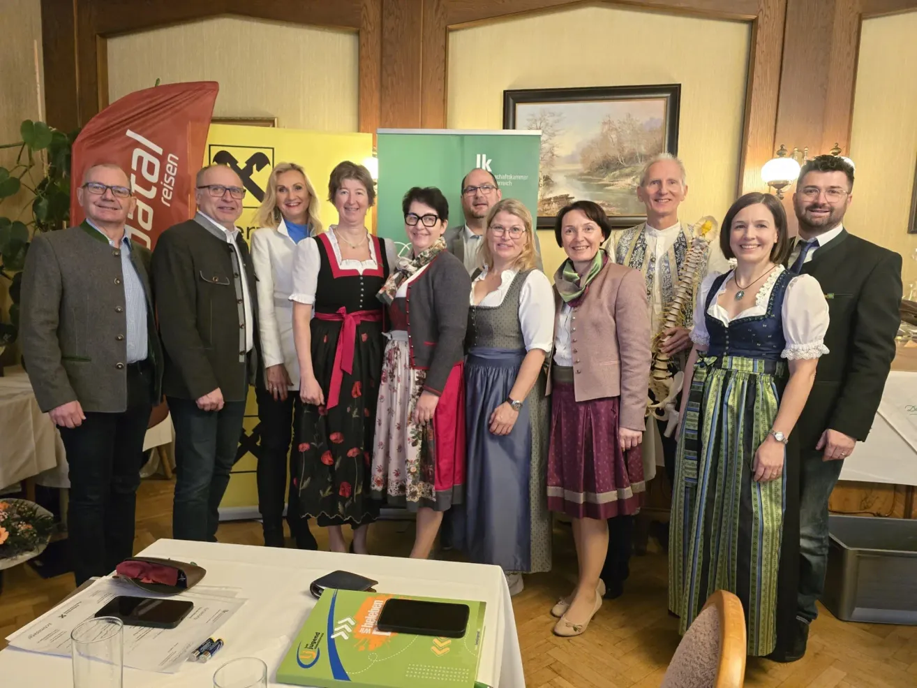 Eine Gruppe von Menschen in traditioneller Kleidung steht gemeinsam in einem Raum und lächelt für ein Foto. Ein Tisch mit einer grünen Box und anderen Gegenständen steht vor ihnen. Ein Bilderrahmen ist an der Wand montiert und eine Lampe ist an der Wand befestigt.