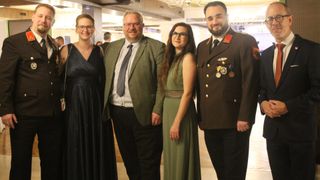 Vier Personen stehen in formeller Kleidung zusammen. Ein Mann in Uniform mit Medaillen, eine Frau in einem grünen Kleid und zwei Männer in Anzügen, einer lächelt, der andere ernst. Sie sind wahrscheinlich bei einer formellen Veranstaltung.