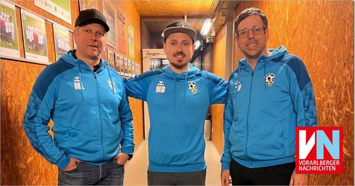 Drei Männer in blauen Kapuzenjacken stehen in einem Flur, lächeln und posieren für ein Foto. Der Mann in der Mitte hat ein Fußballlogo auf seiner Jacke.