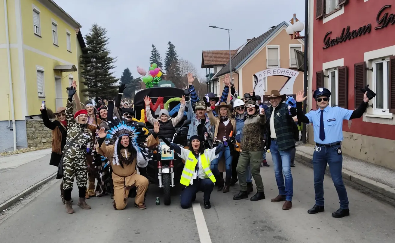 Eine Gruppe von Menschen in verschiedenen Kostümen, darunter ein indianischer Federkopfschmuck, Cowboyhüte und Camouflage, versammelt sich auf einer Straße, einige knien, einige stehen und eine Person sitzt auf einem Motorrad. Hinter ihnen befinden sich Häuser, eine Straßenlaterne und ein Schild, das 'Friedheim' steht.