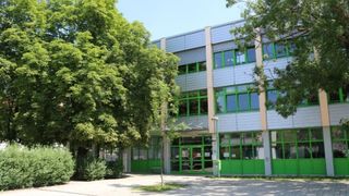 Die Vorderseite eines modernen Gebäudes mit grünen Türen und großen Fenstern. Bäume und Pflanzen umgeben das Gebäude.