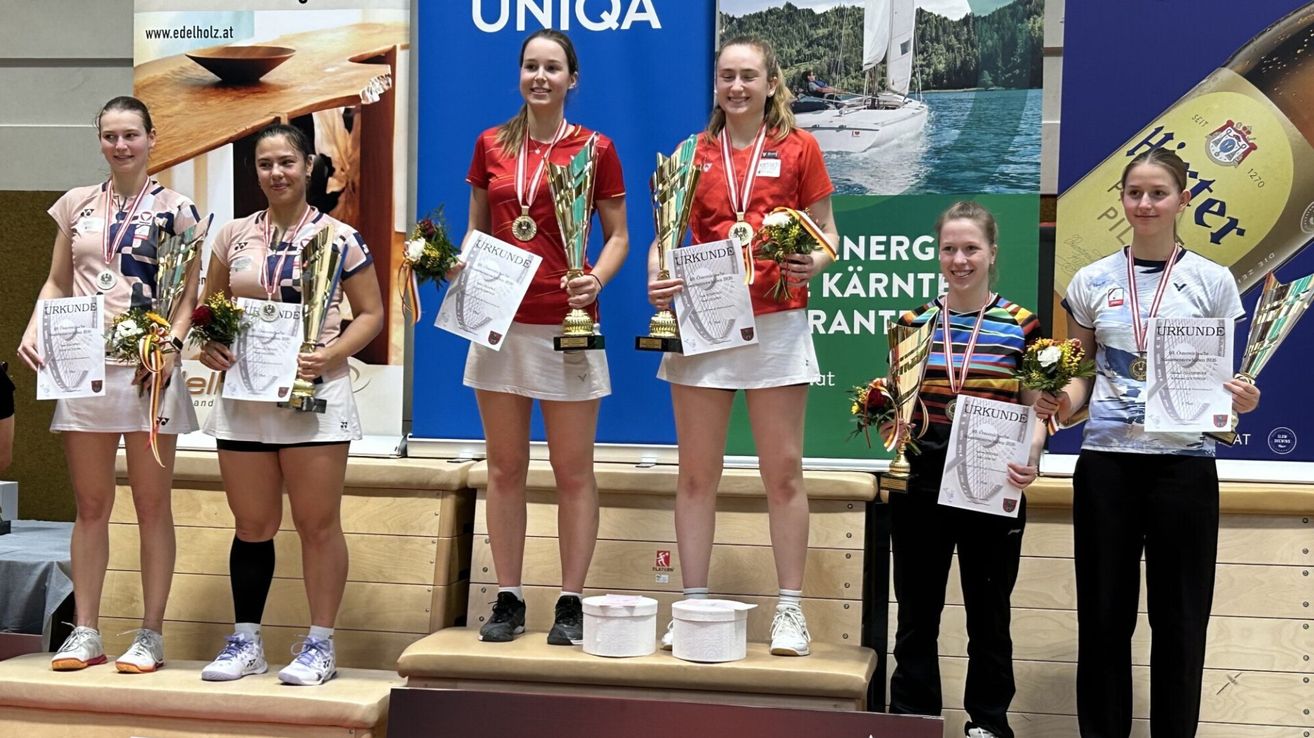 Vier junge Frauen stehen auf einem Podest, jede hält Trophäen und Urkunden. Sie tragen rote Shirts und weiße Röcke. Der Hintergrund zeigt einen blauen Banner mit dem Wort 'UNIQA' und einen grünen Banner mit einem weißen Segelboot und Text.