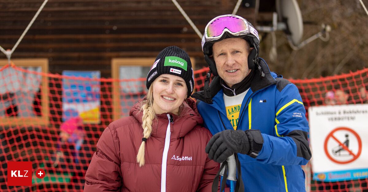 Eine lächelnde Frau und ein Mann in Skiausrüstung stehen gemeinsam in einer verschneiten Umgebung, mit einem bunten Hindernis dahinter.