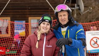 Eine lächelnde Frau und ein Mann in Skiausrüstung stehen gemeinsam in einer verschneiten Umgebung, mit einem bunten Hindernis dahinter.