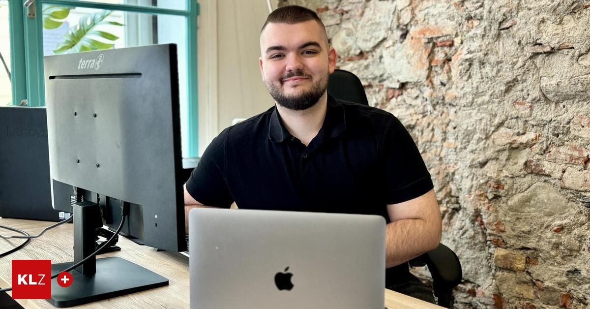 Ein Mann sitzt an einem Schreibtisch mit einem Computerbildschirm und einem Laptop und lächelt in einem Büro mit einer Backsteinmauer.