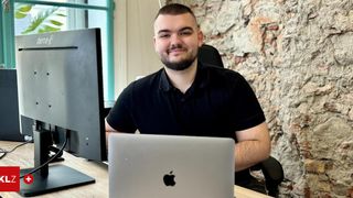 Ein Mann sitzt an einem Schreibtisch mit einem Computerbildschirm und einem Laptop und lächelt in einem Büro mit einer Backsteinmauer.