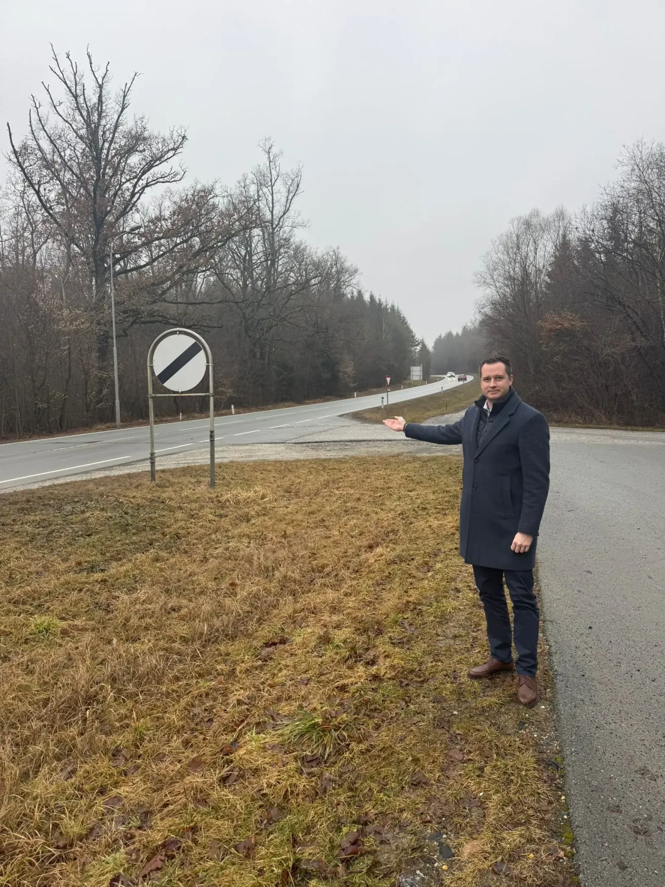 Ein Mann in einem dunklen Mantel steht am Straßenrand und zeigt mit der Hand. Ein Schild mit einem schwarzen Streifen steht hinter ihm.