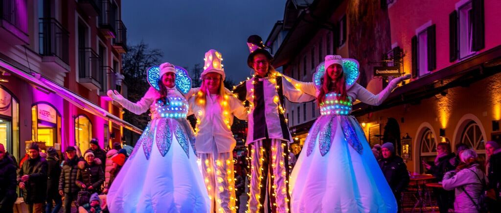 Drei Menschen in LED-Kostümen gehen bei Nacht auf Stelzen, während eine Menge von Menschen zuschaut. Die Straße ist mit Lichtern geschmückt. Hinter ihnen befinden sich Gebäude mit Vordächern und einem Schild.