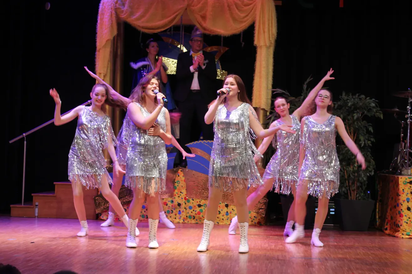 Eine Gruppe von Frauen in Paillettenkleidern und weißen Stiefeln tritt auf der Bühne auf, zwei halten Mikrofone und lächeln. Dahinter klatschen ein Mann im Anzug und eine Frau in einem blauen Kleid.