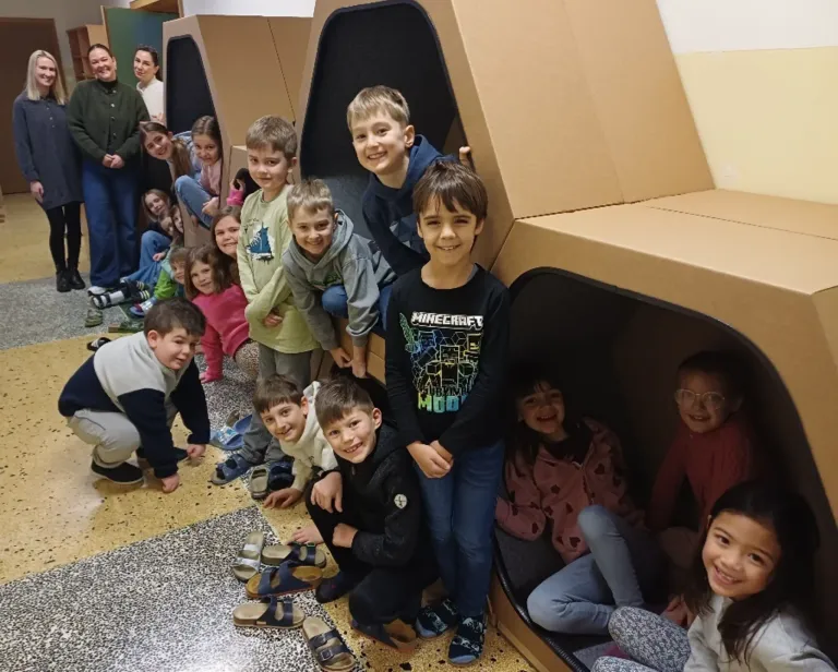 A group of children and adults are posing inside a cardboard structure with shoes and slippers scattered on the floor.