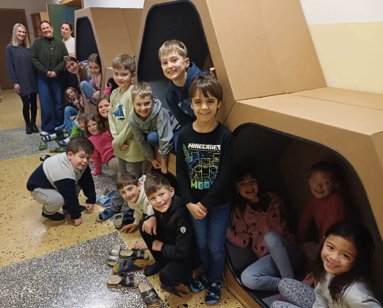 A group of children and adults are posing inside a cardboard structure with shoes and slippers scattered on the floor.