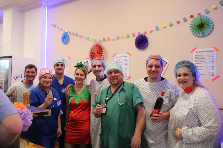 A group of people dressed in colorful costumes and medical attire are posing for a photo. A woman in a strawberry costume holds a glass, while a man in a green robe holds a bottle. The background features a colorful garland and a paper fan.