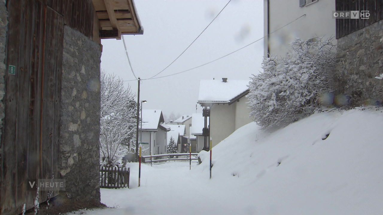 Eine verschneite Straße mit Häusern und Bäumen, die in Schnee eingehüllt sind. Versorgungsmasten und Drähte sind oben sichtbar. Ein kleiner Zaun und ein Laternenpfahl sind vorhanden.