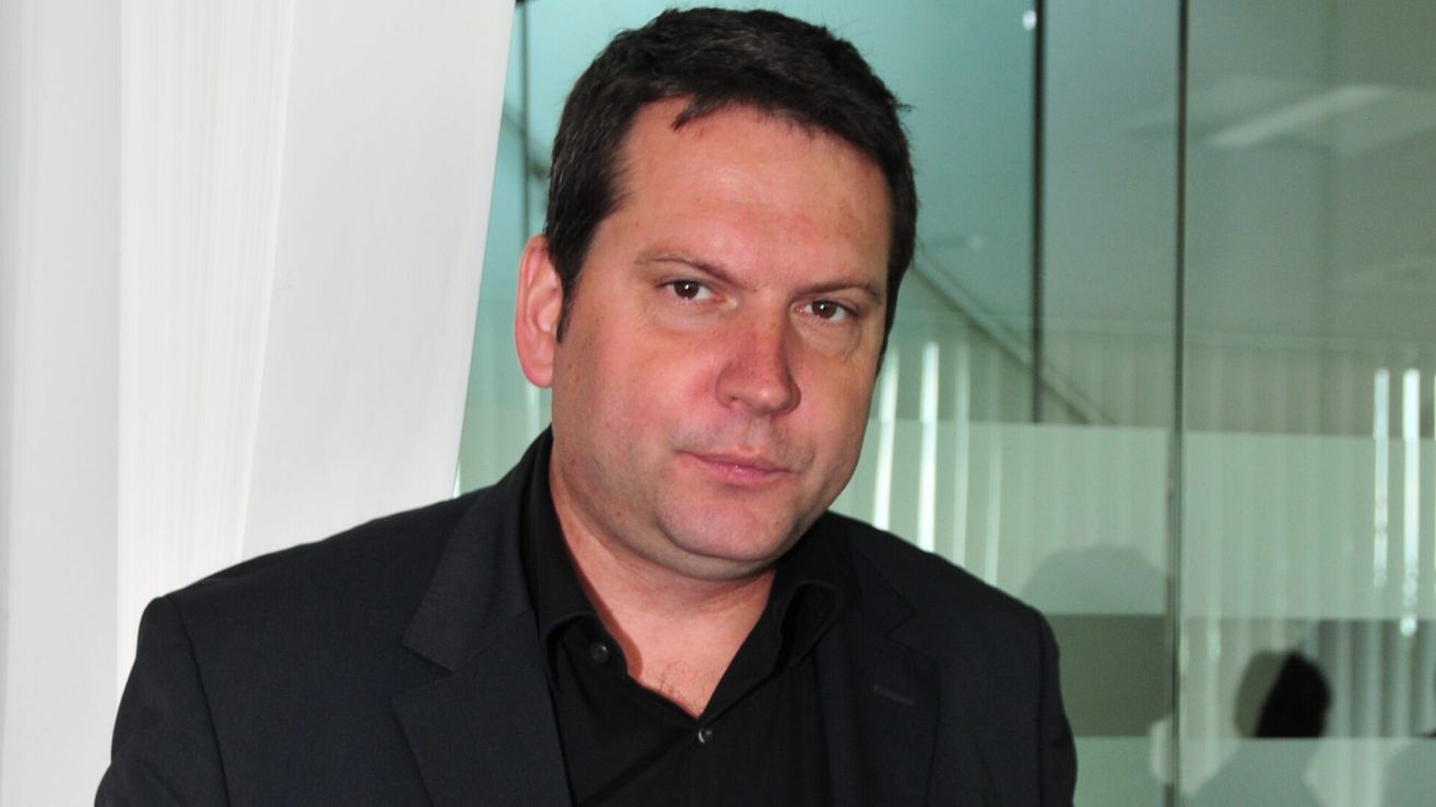 Ein Mann mit kurzem schwarzen Haar trägt ein schwarzes Hemd und sieht nach vorne. Er scheint für ein Foto zu posieren. Hinter ihm ist ein unscharfer Hintergrund aus einer weißen Wand und einer grünen Glaswand.