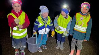 Fünf Kinder in Sicherheitswesten und Stiefeln stehen nachts auf einem Schotterweg. Ein Kind hält einen durchsichtigen Eimer. Sie tragen alle Mützen, Handschuhe und Stirnlampen.