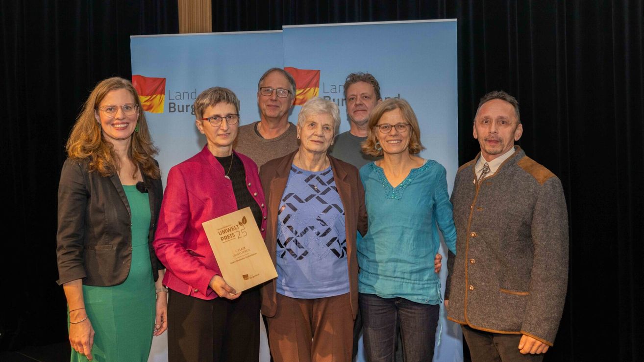 A group of individuals stand together holding an award. A woman holds a certificate with a green leaf symbol. Behind them is a backdrop with the text 'Land Burg'.