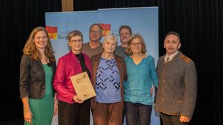 Eine Gruppe von Personen steht zusammen und hält eine Auszeichnung. Eine Frau hält ein Zertifikat mit einem grünen Blattsymbol. Hinter ihnen ist ein Hintergrund mit dem Text 'Land Burg'.