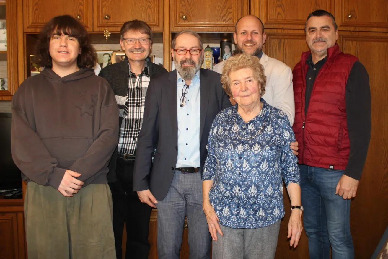 Eine Gruppe von Menschen posiert für ein Foto in einer Küche. Eine Frau in einem Blumenhemd steht vorne mit einem Lächeln. Hinter ihr stehen drei Männer. Einer von ihnen trägt eine Jacke und eine Brille. Hinter ihnen steht ein Holzschrank.