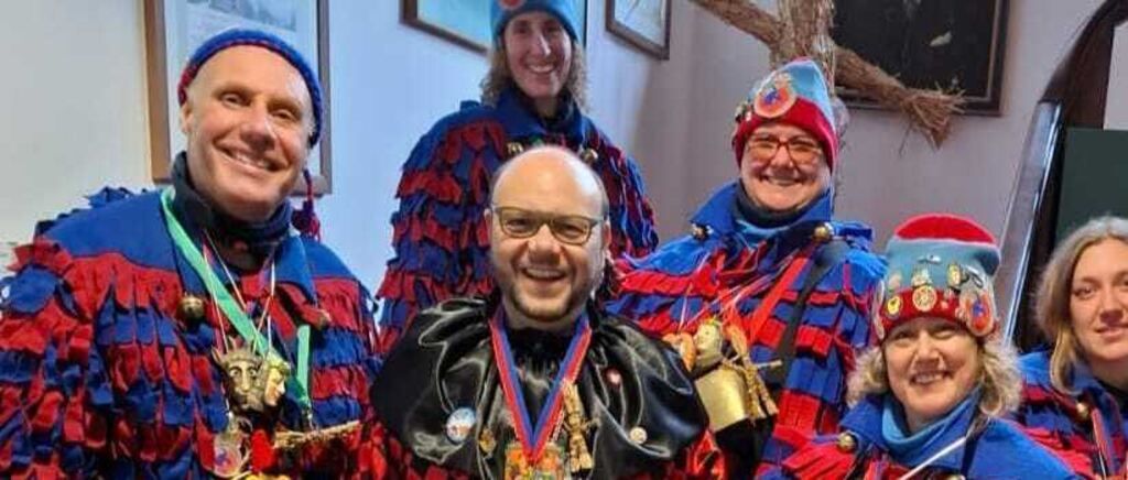 Sechs Menschen in festlicher Kleidung, lächelnd, stehen zusammen. Drei tragen Rot und Blau, zwei tragen Blau und Rot. Ein Mann in Schwarz und Rot trägt eine Brille und eine Halskette.