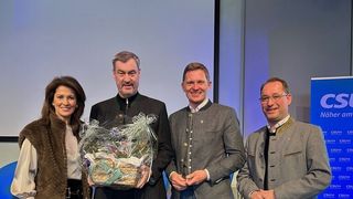 Vier Personen, drei Männer und eine Frau, stehen zusammen und lächeln für ein Foto, halten einen Geschenkkorb mit einer Schleife. Sie sind in traditioneller deutscher Kleidung gekleidet.