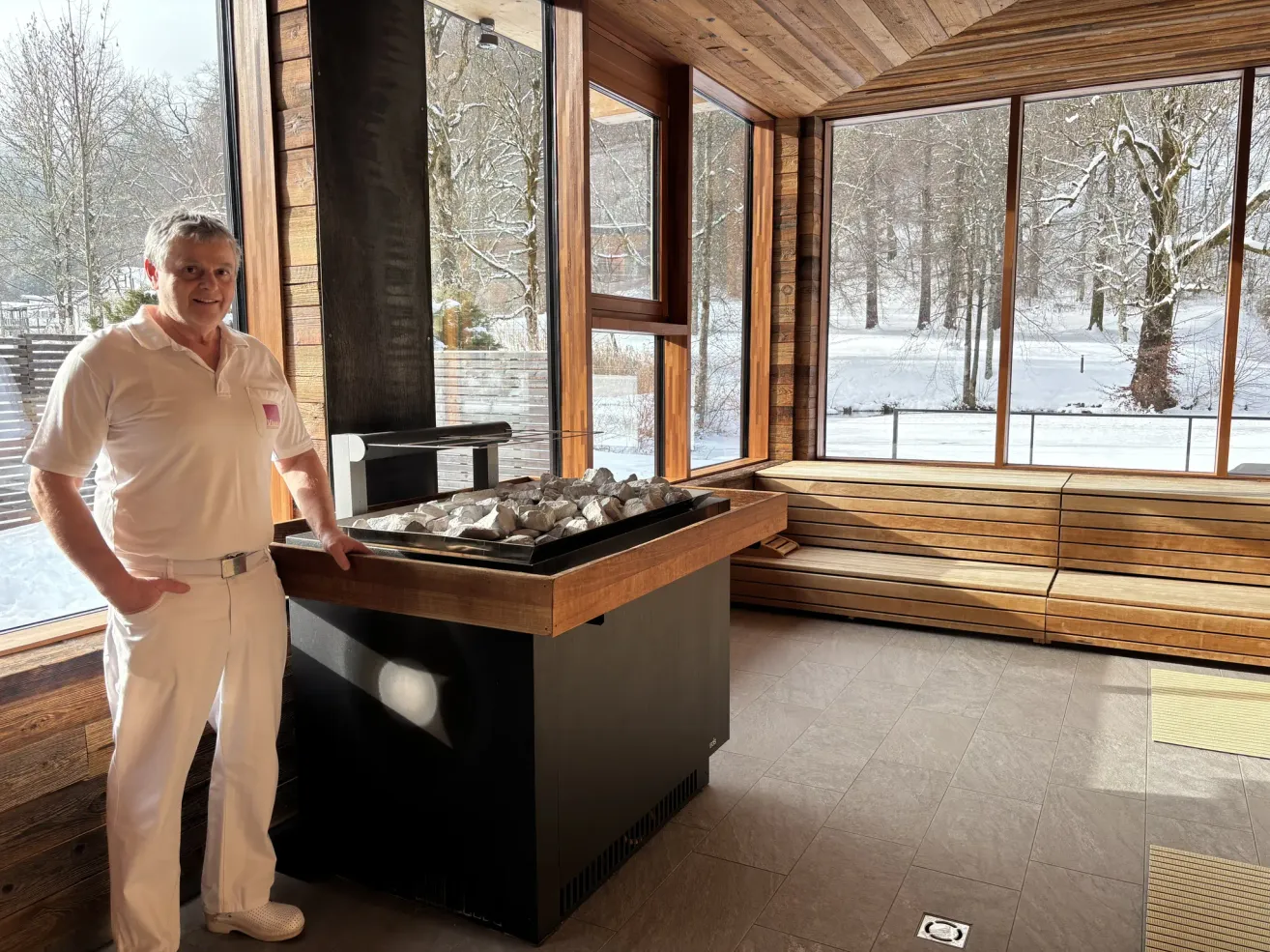 Ein Mann in Weiß steht in einem Raum mit einer Holzbank, einem Kamin und großen Fenstern, die auf eine verschneite Landschaft hinausblicken.