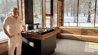 Ein Mann in Weiß steht in einem Raum mit einer Holzbank, einem Kamin und großen Fenstern, die auf eine verschneite Landschaft hinausblicken.