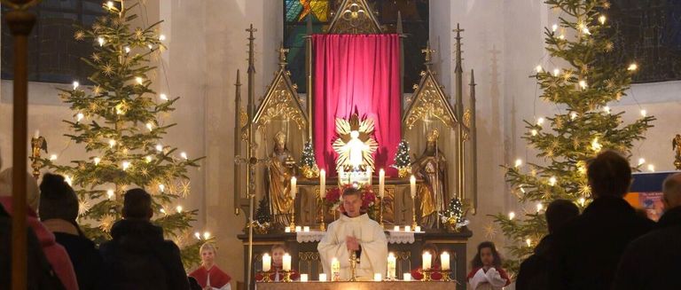 Ein Priester steht vor einem Altar, umgeben von brennenden Kerzen, während Kinder und Erwachsene in einer mit Weihnachtsbäumen und einem roten Vorhang geschmückten Kirche zusehen.