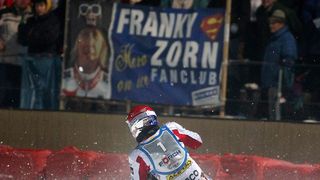 Ein Skifahrer in einem roten und weißen Rennanzug mit der Nummer 1 darauf fällt im Schnee. Dahinter befindet sich ein Schild mit der Aufschrift Franky Zorn Fanclub und einem Superman-Logo.
