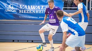 In einer Sporthalle drinnen, ein Spieler in einem lila Trikot dribbelt einen Fußball, während zwei Gegner in blauen Trikots versuchen, ihn zu blockieren.