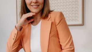 Eine Frau in einem pfirsichfarbenen Blazer und weißem Shirt sitzt lächelnd. Sie hat eine Hand am Kinn und sitzt an einer weißen Wand.