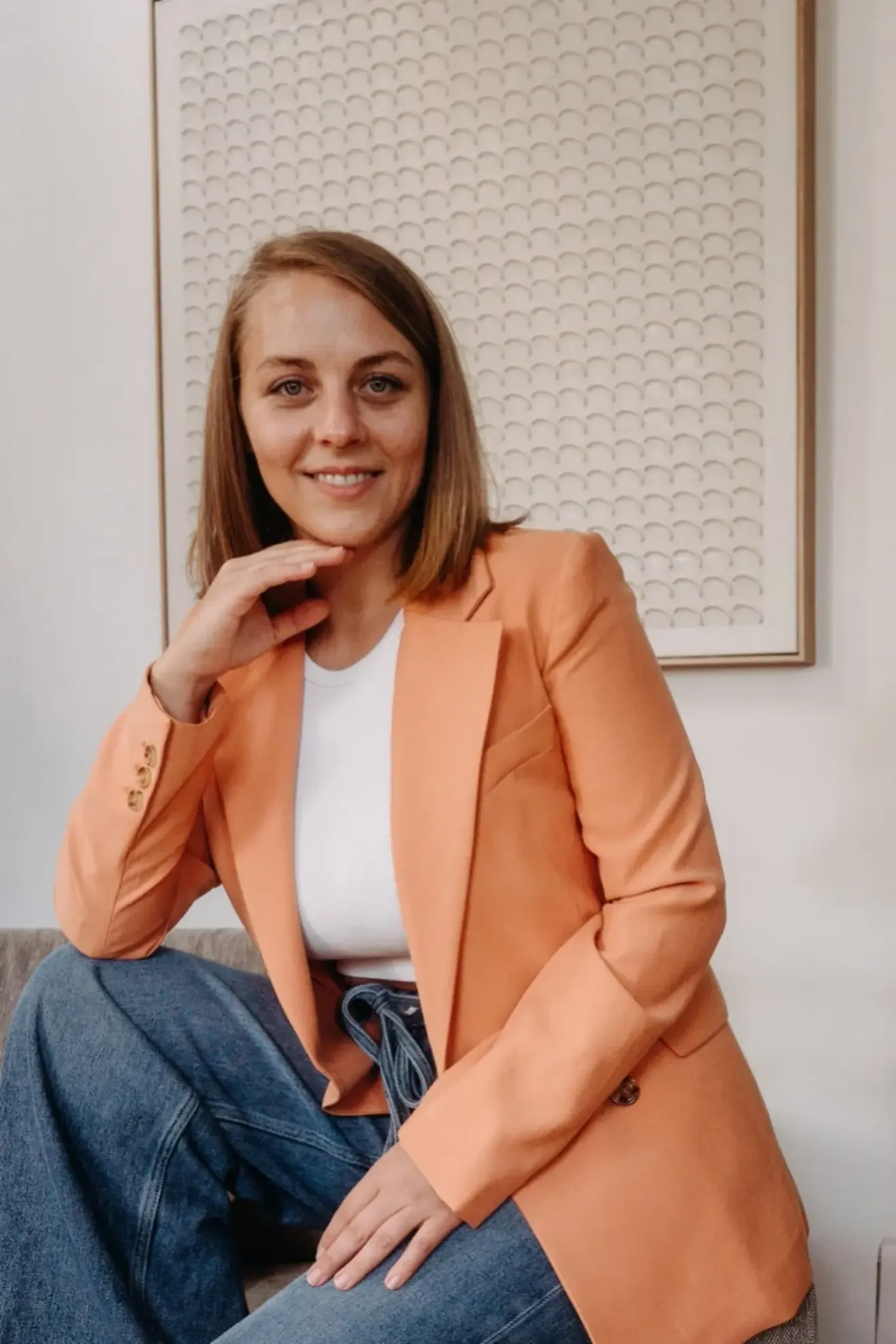 Eine Frau in einem pfirsichfarbenen Blazer und weißem Shirt sitzt lächelnd. Sie hat eine Hand am Kinn und sitzt an einer weißen Wand.