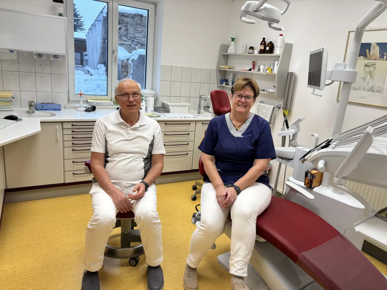 Ein älterer Mann und eine ältere Frau sitzen in einer Zahnarztpraxis. Sie tragen beide eine Brille und lächeln in die Kamera. Hinter ihnen sind Zahnarztgeräte und ein Waschbecken zu sehen.