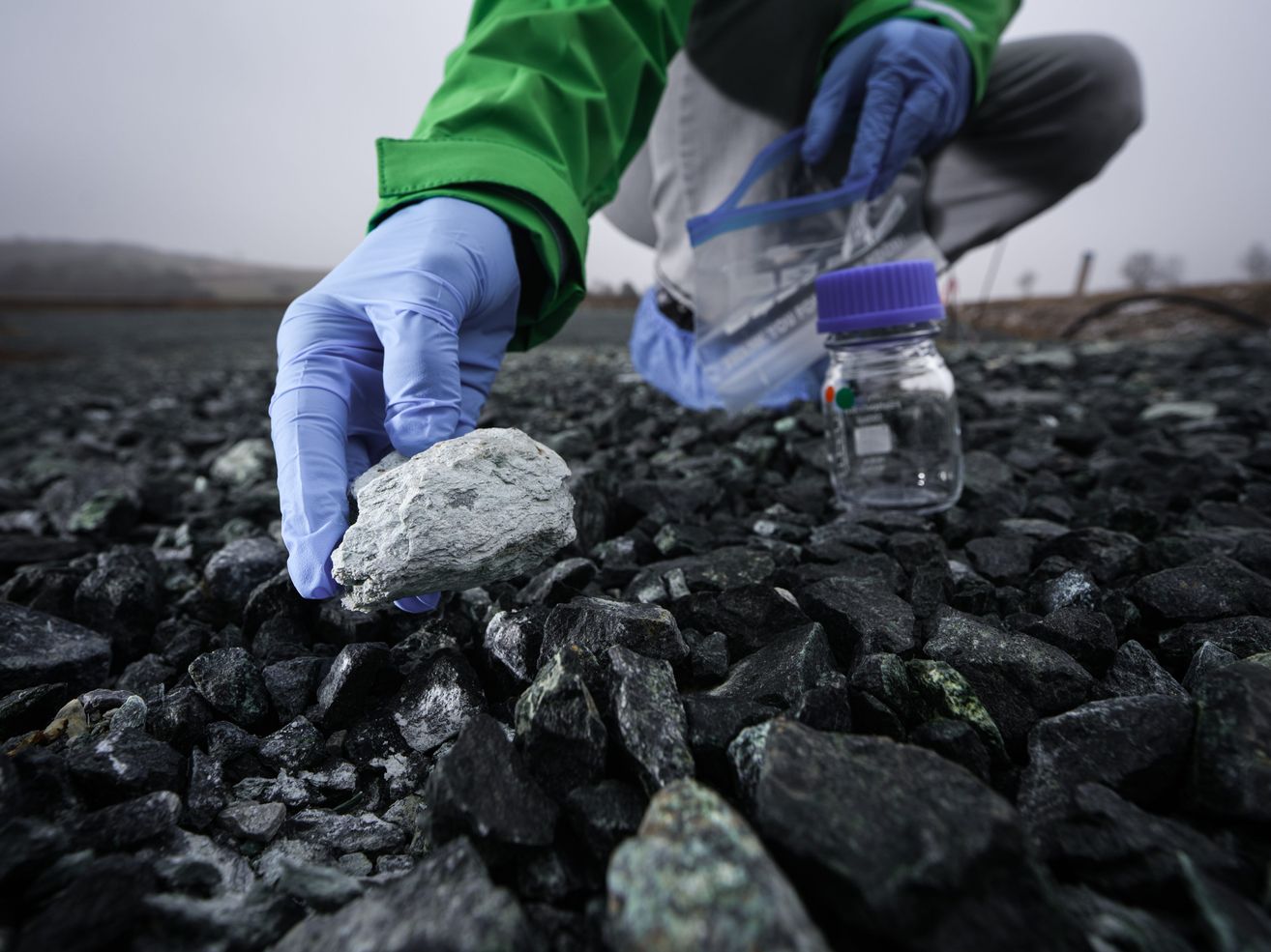 Eine Person in einem grünen Jacke und blauen Handschuhen hält einen Stein und ein Glas mit einem lila Deckel auf einer felsigen Oberfläche.