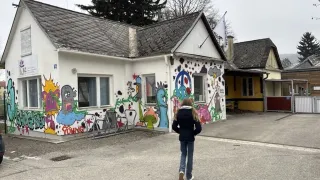Ein junges Mädchen geht an einem weißen Haus mit bunten Graffiti, geparkten Fahrrädern davor und Bäumen im Hintergrund vorbei.