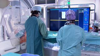 Zwei medizinische Fachkräfte in Operationskleidung arbeiten in einem Krankenhauszimmer, eine beobachtet einen Monitor und die andere bereitet sich möglicherweise auf eine Operation vor.