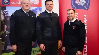 Drei Männer in Uniformen stehen zusammen vor einem roten Hintergrund mit Text und lächeln für ein Foto.