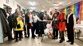 Eine Gruppe von Menschen in Kostümen, darunter Mönche und ein Priester, posiert um einen dekorierten Tisch mit Luftballons. Sie lächeln und scheinen eine gute Zeit zu haben.