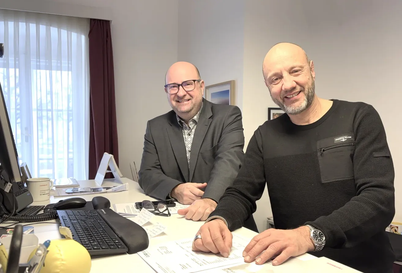 Zwei Männer sitzen in einem Büro an einem Schreibtisch, lächeln und betrachten ein Dokument. Der Mann auf der linken Seite trägt eine Brille und einen Anzug, während der Mann auf der rechten Seite ein schwarzes Shirt und eine Uhr trägt.