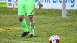 Ein Fußballtorwart in einer grünen Uniform steht auf einem Feld mit einem Fußball vor ihm. Der Spieler ist schlammig und trägt Handschuhe und schwarze Schuhe.