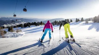 Zwei Skifahrer rutschen einen verschneiten Hang hinunter. Einer trägt eine pinke Jacke, blaue Hosen und schwarze Handschuhe, der andere gelbe Hosen und schwarze Handschuhe. Im Hintergrund befindet sich eine Gondelbahn.