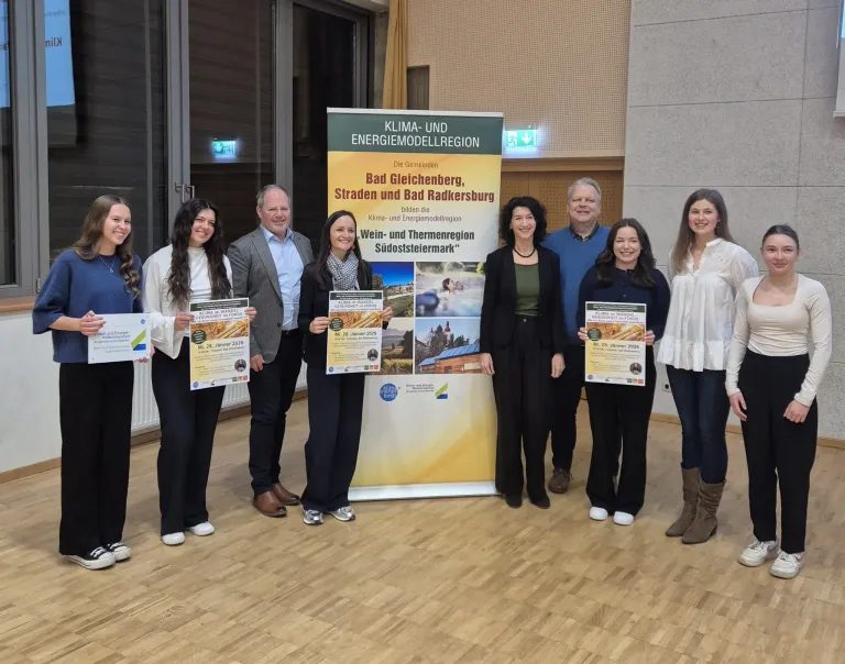 A group of people stands in front of a banner with a poster in their hands. They are smiling and posing for a photo. Behind them, a glass wall reflects the surroundings.