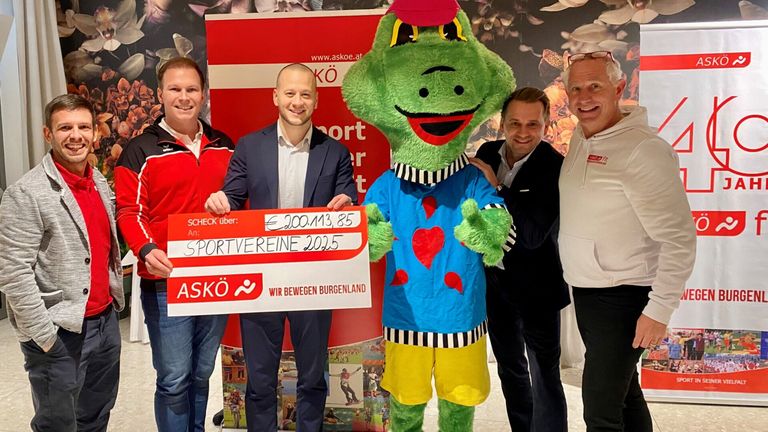 Fünf Männer posieren mit einem Maskottchen. Ein Mann hält einen großen Scheck, das Maskottchen trägt ein Kostüm mit Herz und Streifen. Der Scheck ist für einen Sportverein.