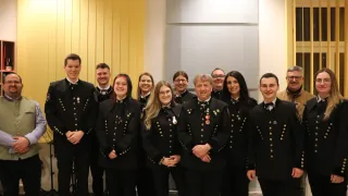 Eine Gruppe uniformierter Personen steht zusammen in einem Raum und lächelt für ein Foto. Sie sind in schwarze Uniformen mit goldenen Knöpfen und Medaillen gekleidet. Hinter ihnen befindet sich eine gelbe Wand und eine graue quadratische Struktur.